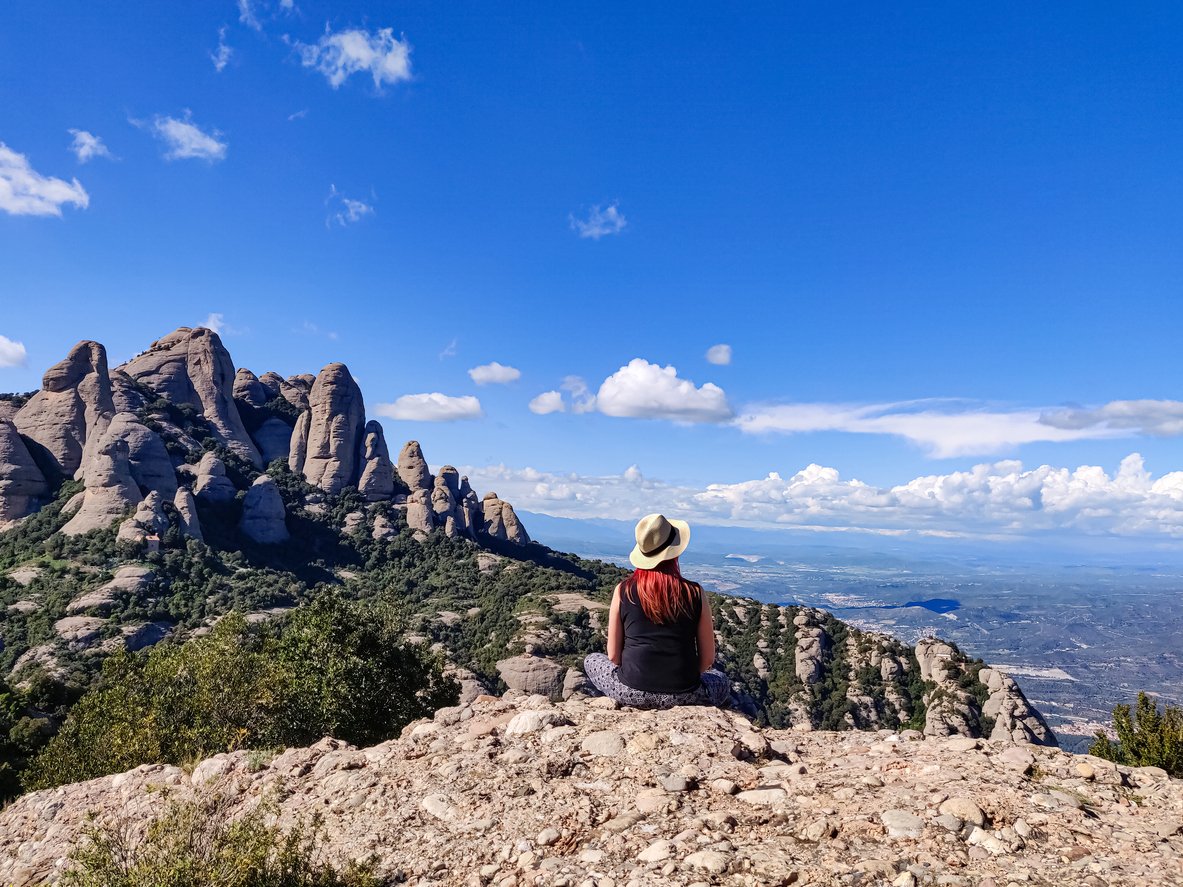 A woman wearing a hat sits on a rocky outcrop overlooking the Montserrat Mountains near Barcelona, with dramatic rock formations and a vast landscape stretching into the horizon under a bright blue sky.