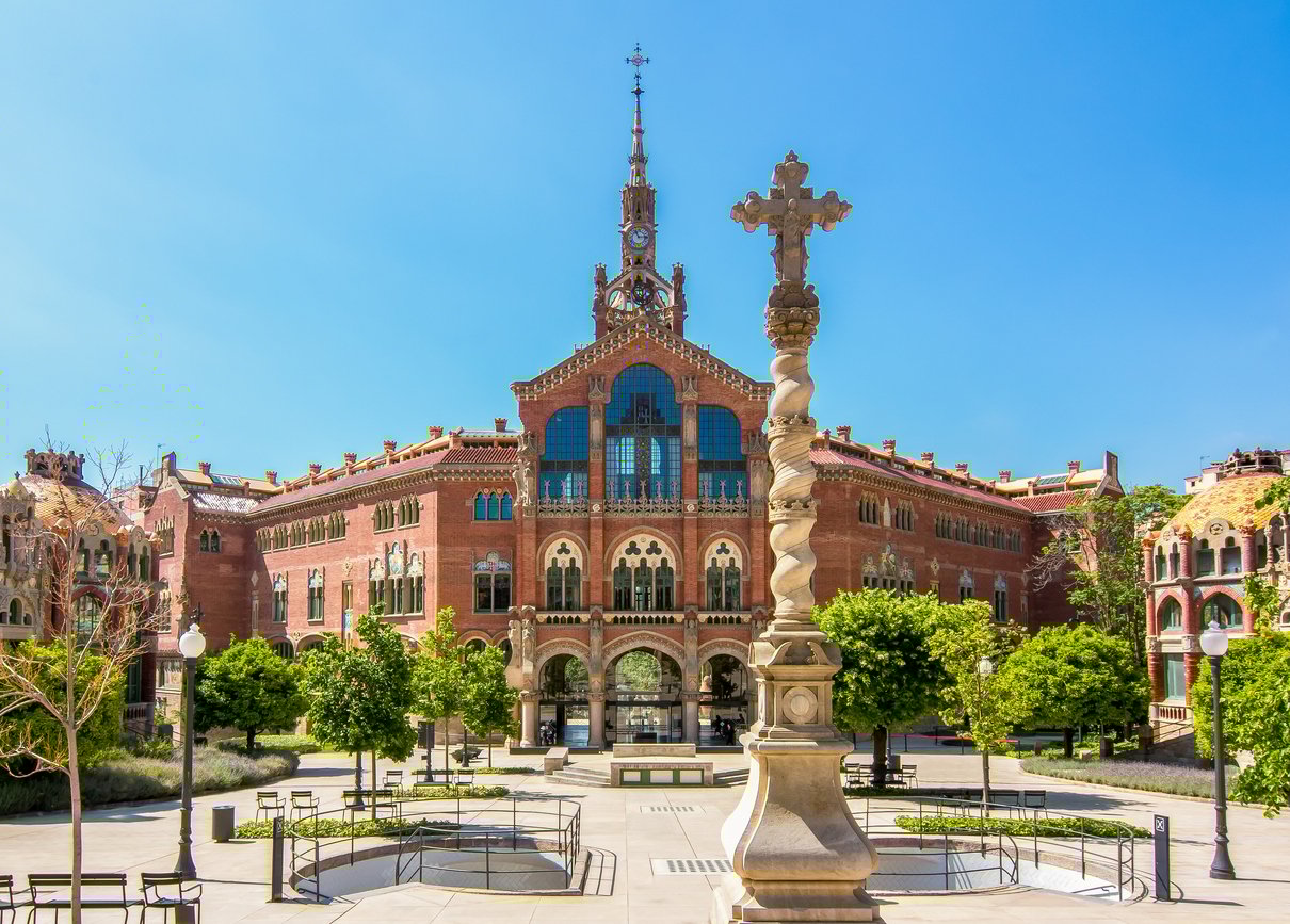 Sant Pau Hospital, a stunning example of modernist architecture in Barcelona, featuring red brick buildings with ornate facades, colorful tiles, and a central spire, surrounded by a peaceful garden courtyard.