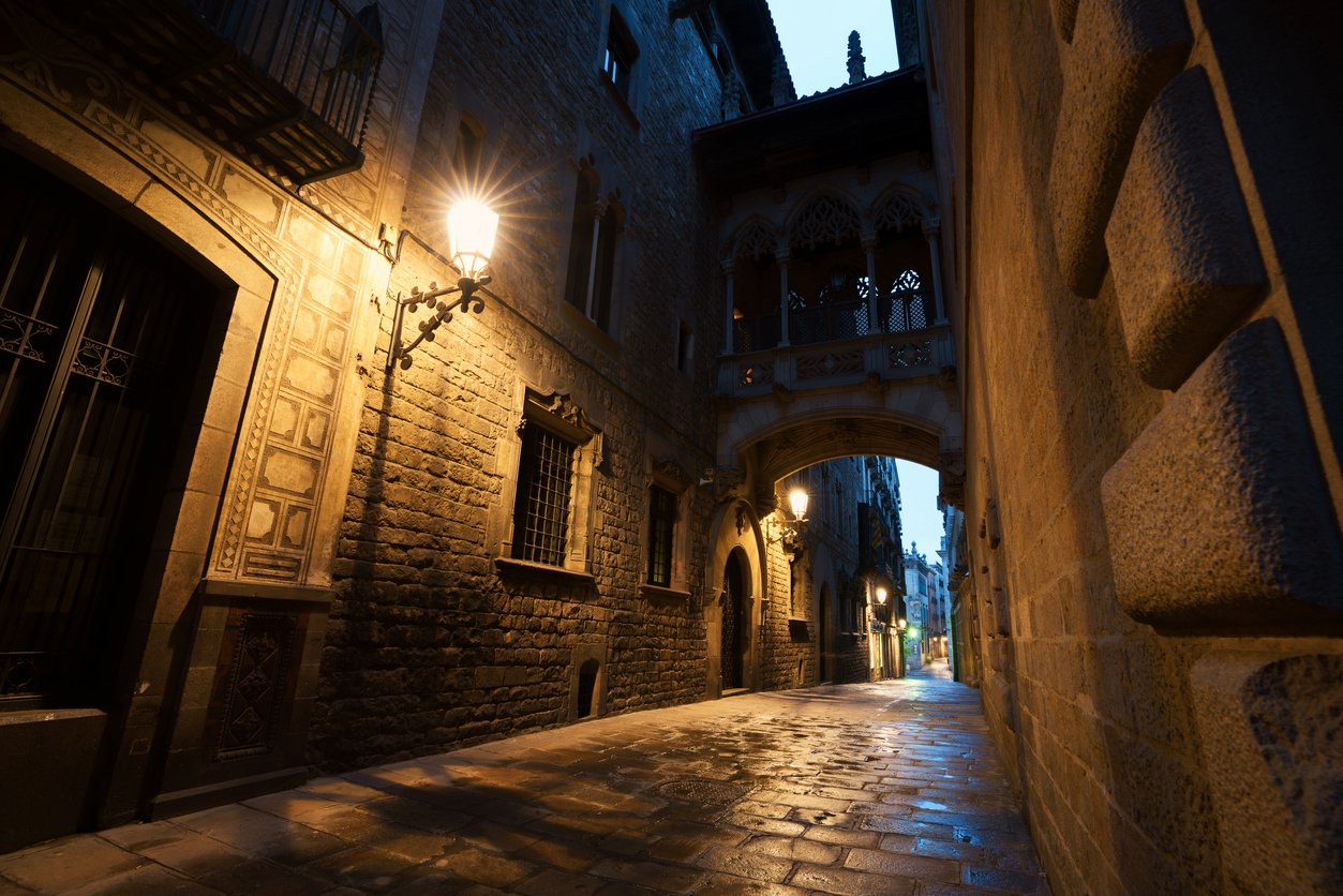 Dimly lit narrow street in the Gothic Quarter of Barcelona at night, featuring an archway and historic stone buildings."