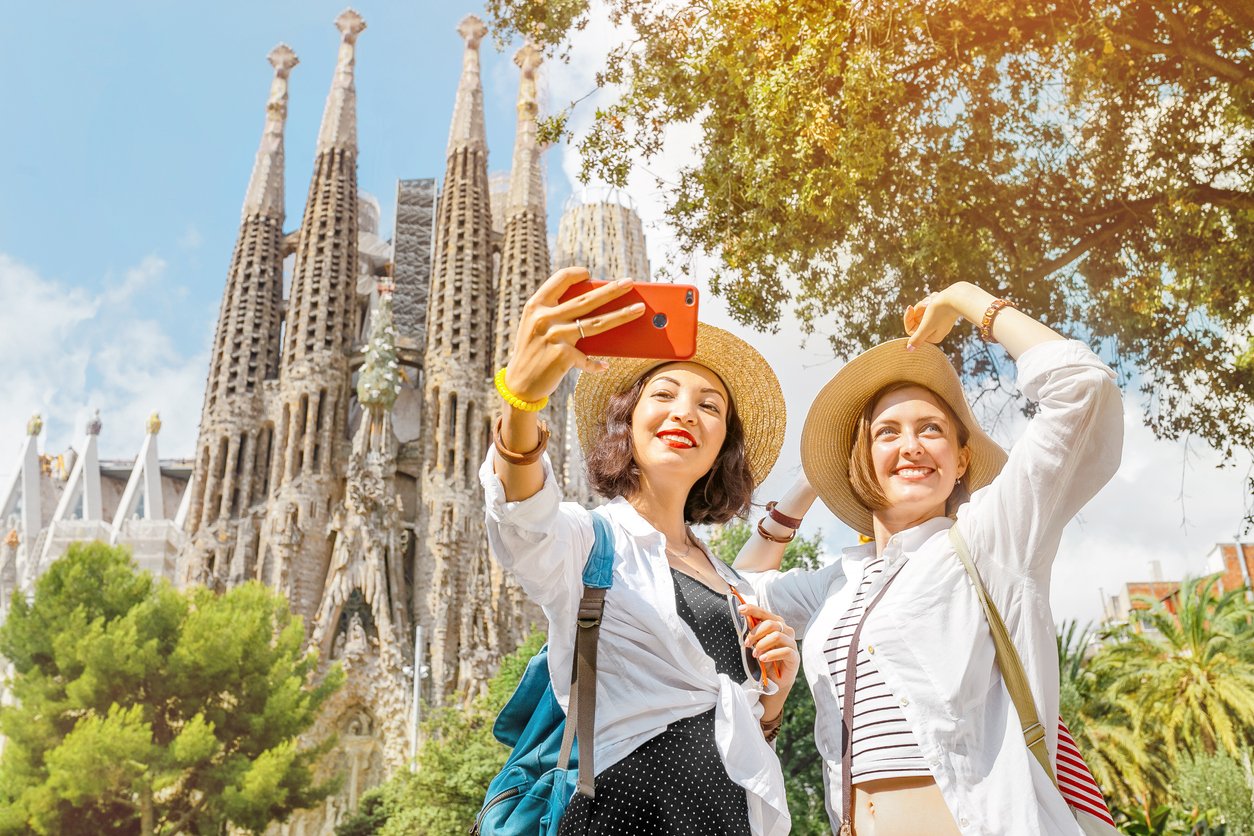 Two women taking a selfie in front of La Sagrada Familia in Barcelona on a sunny day.