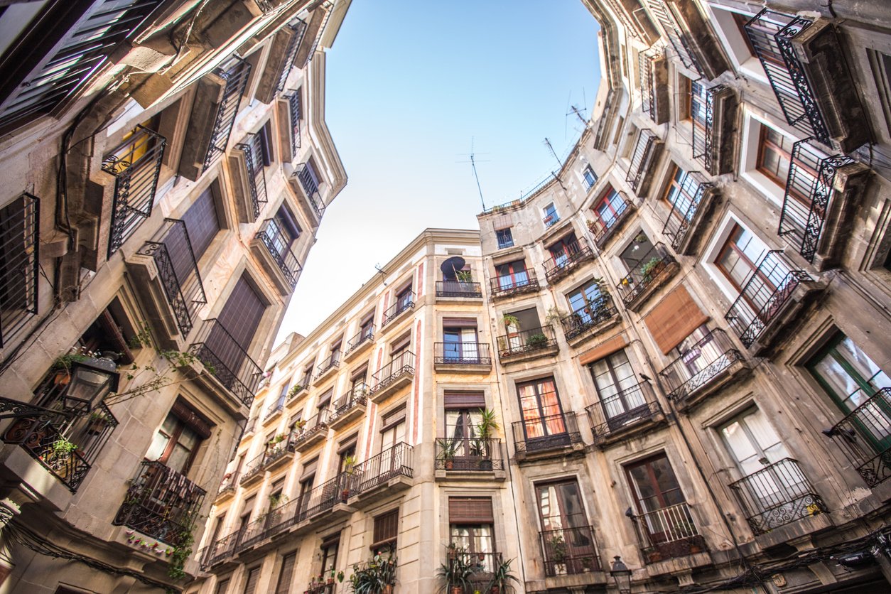 Upward view of residential buildings in Barcelona's Old Town with intricate balconies and plants, bathed in natural light under a clear blue sky.