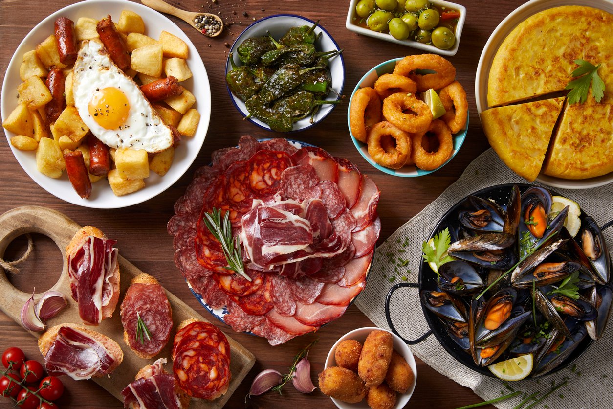 A delicious spread of traditional Spanish tapas, including patatas bravas with a fried egg, chorizo, pimientos de padrón, green olives, calamari rings, tortilla española, cured meats, mussels in a black skillet, croquettes, and jamón ibérico on bread.
