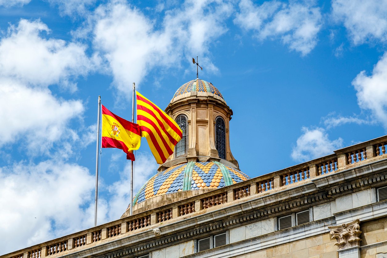 Spanish and Catalan flags on top of a government building in Barcelona, waving under a blue sky.