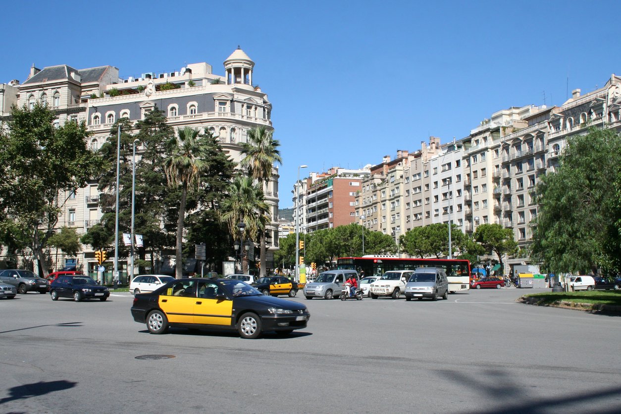 Busy intersection in Barcelona, featuring classic yellow-and-black taxis, cars, and buses surrounded by elegant buildings and palm trees under a clear blue sky.