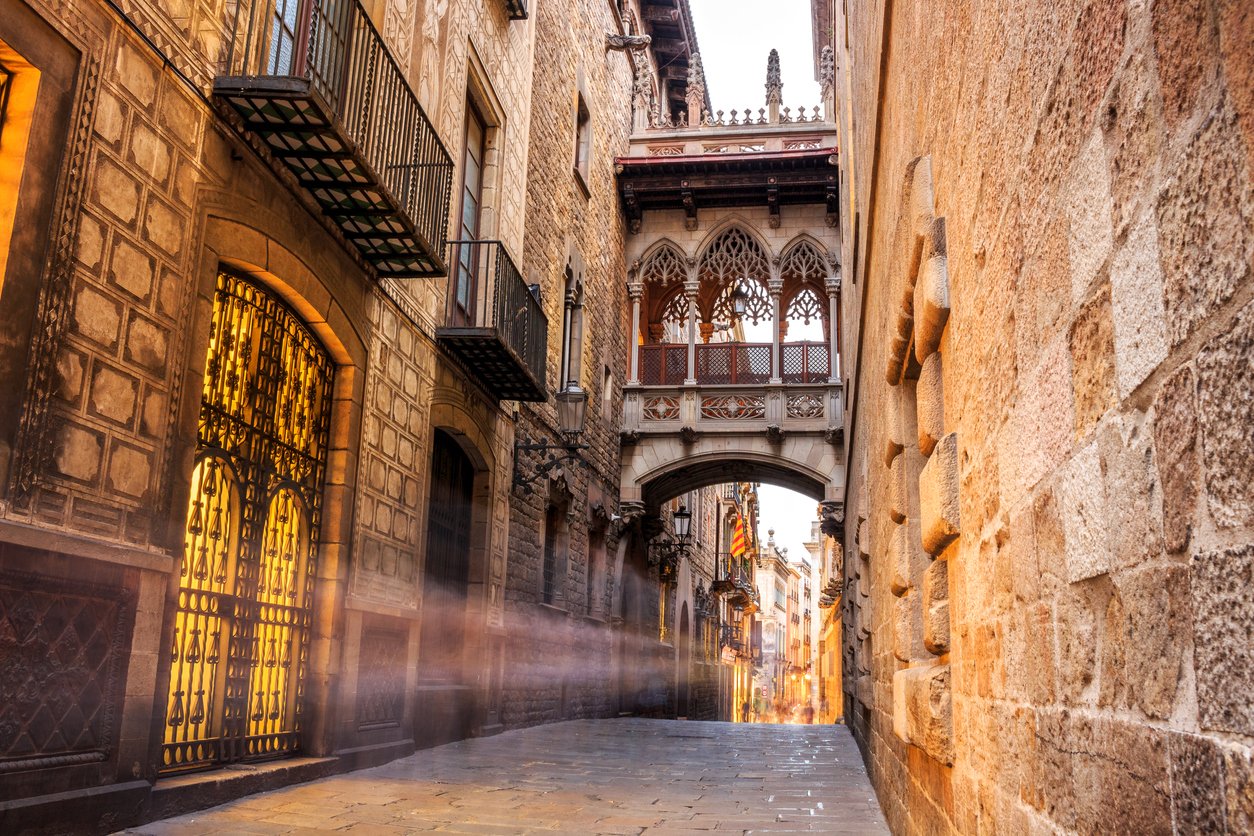 View of the Gothic Quarter in Barcelona featuring the iconic Pont del Bisbe bridge and historic stone buildings lit by warm light.