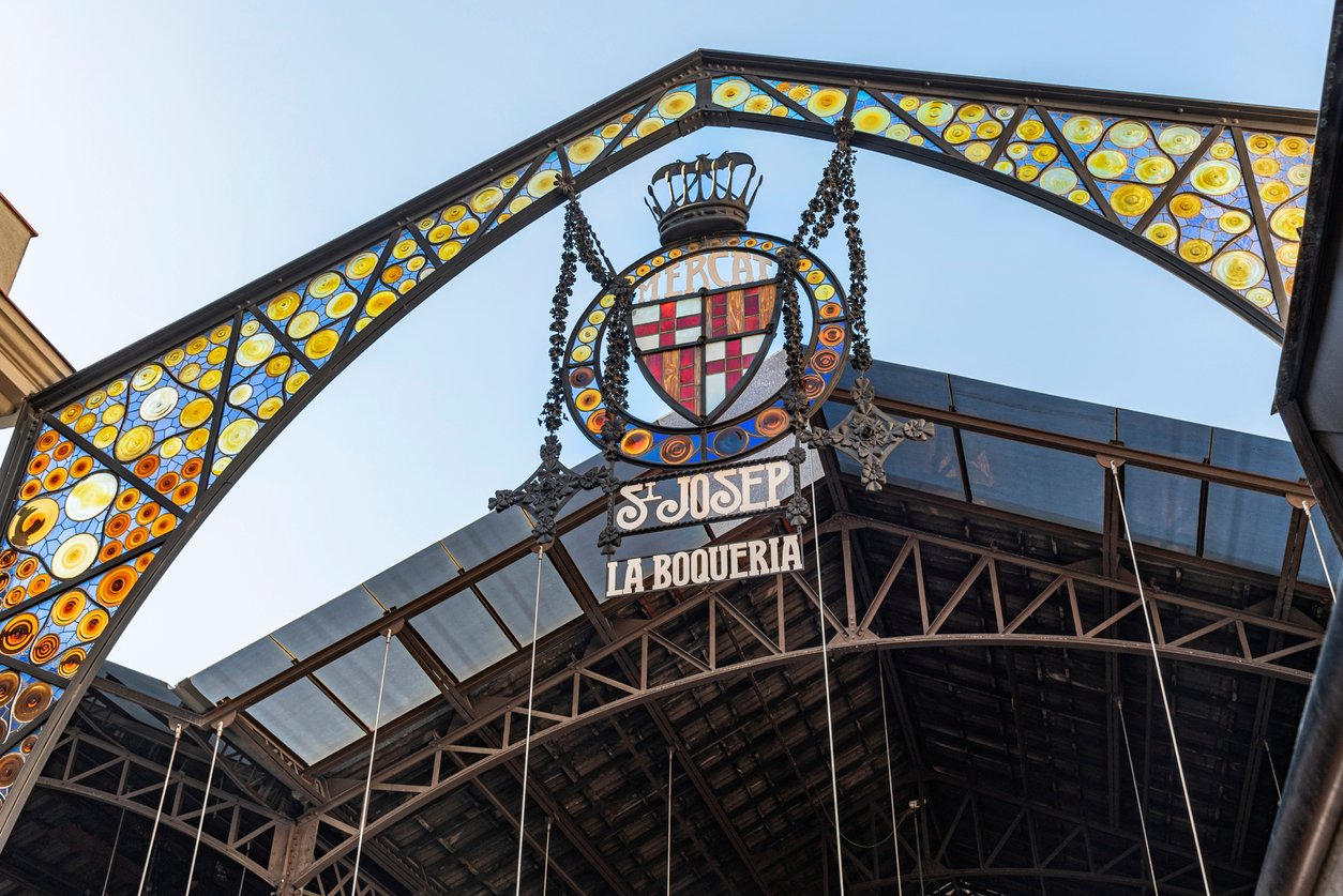 The colorful stained-glass entrance of La Boqueria Market in Barcelona, featuring the sign "Mercat St. Josep La Boqueria" with a vibrant display of circular patterns and ironwork under a clear sky.