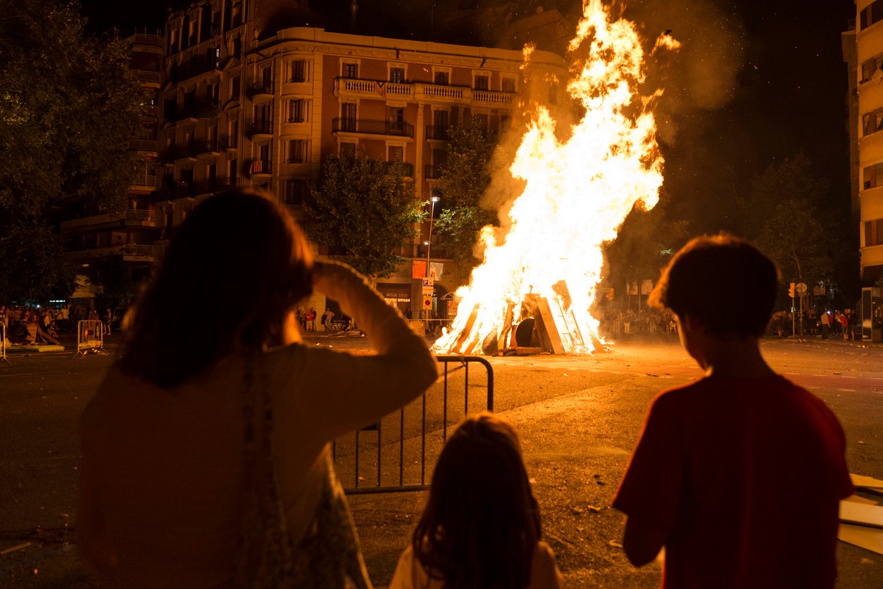 Large bonfire during a nighttime city festival, with spectators watching in a lively urban setting surrounded by residential buildings.
