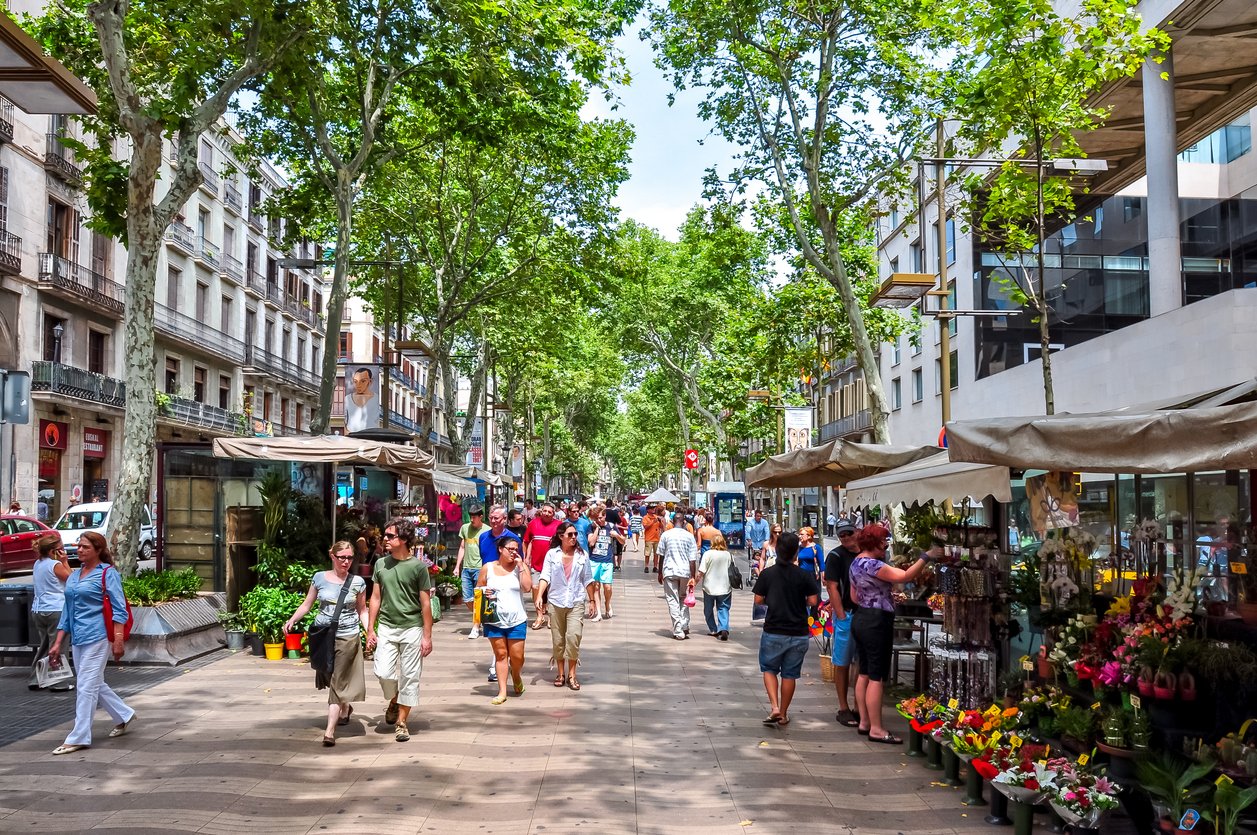 Busy scene of La Rambla in Barcelona, with people strolling past market stalls and lush trees lining the walkway.