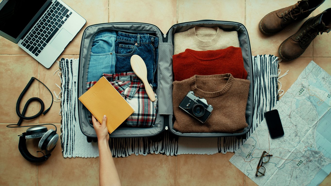 Overhead view of an open suitcase packed with clothes, a camera, and a book, surrounded by travel essentials like a laptop, headphones, boots, map, and smartphone, ready for a trip.
