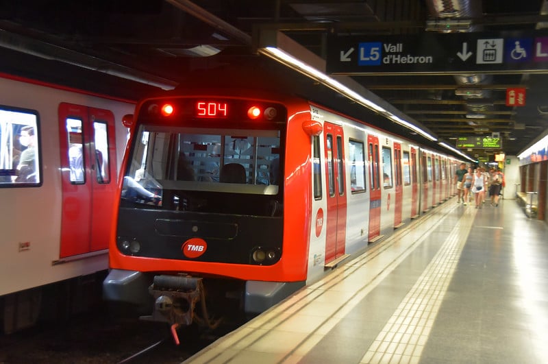A red and white Barcelona Metro train, labeled 504, arrives at a station on the L5 Vall d'Hebron line. The platform is lit with overhead lights, and a few passengers are seen walking on the platform.