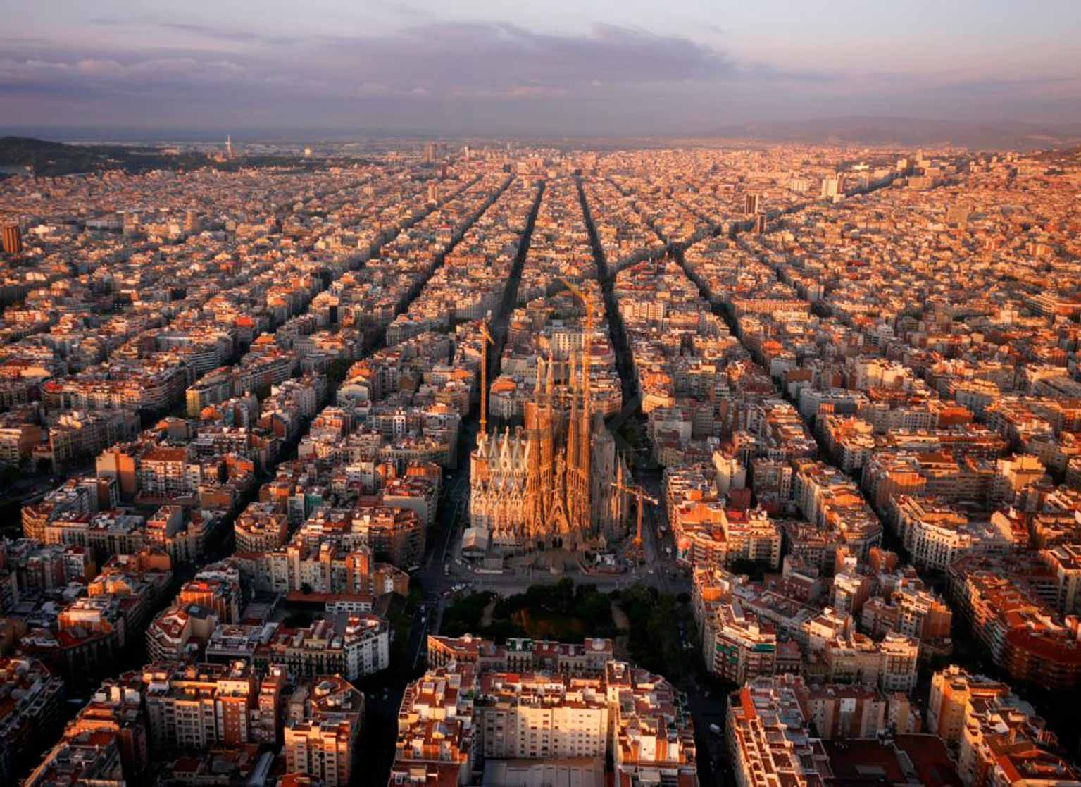 Aerial view of Barcelona's Eixample district at sunset, with the iconic Sagrada Familia cathedral at the center, highlighting the city's grid pattern and urban sprawl.